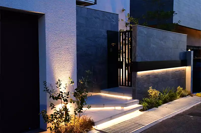 ライトアップされた代理石彫タイルを使用し高級感のあるモダンな外構の門袖と門まわりとフロート階段と植栽のライトアップの写真