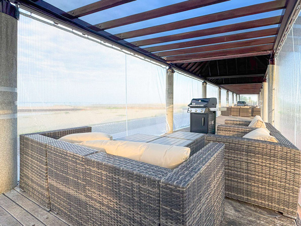 海辺のウッドデッキに連なる木製パーゴラの下、透明ポリカ屋根越しに青空が見え、側面はメッシュの防風スクリーンで囲われ、屋外ソファとBBQグリルが並ぶ屋外BBQ施設の様子