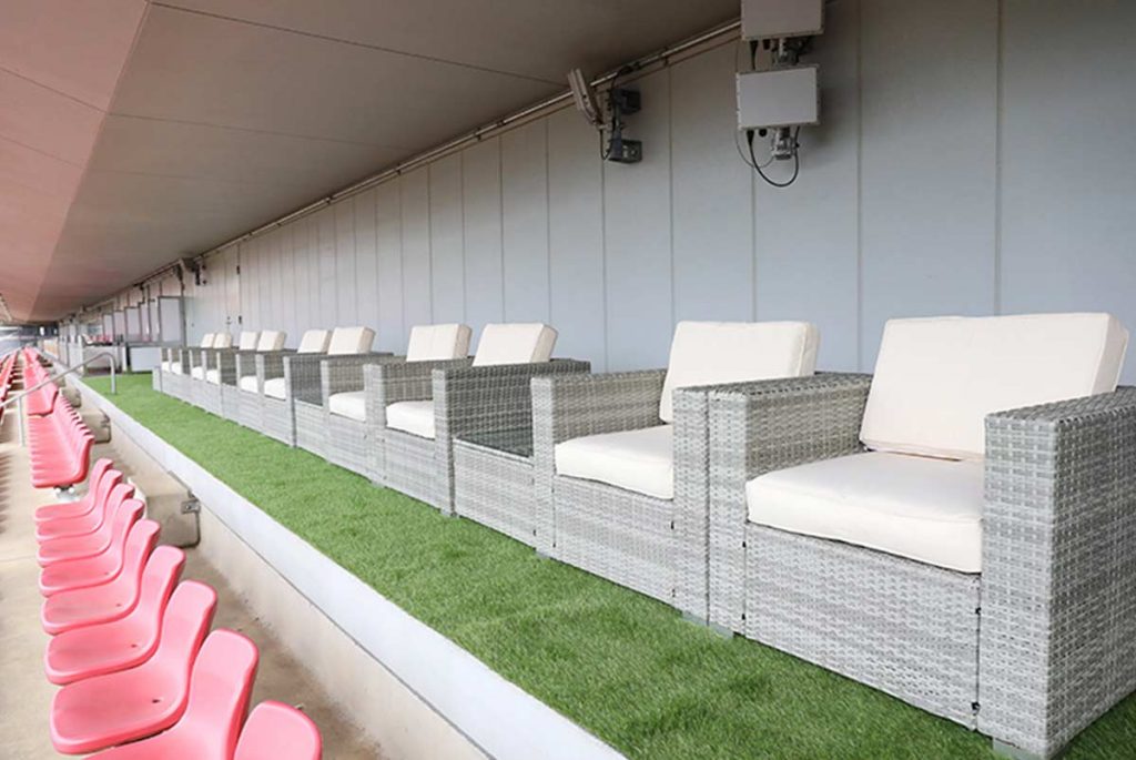 カシマサッカースタジアムのプレミアムソファシート - ラタン素材の高級感あふれるVIP観戦席。白いクッションと灰色のラタン編みがスタイリッシュで、人工芝の上に配置された快適な観戦環境。鹿島アントラーズのホームスタジアムでの特別な観戦体験を提供するバックスタンド側のソファシート。