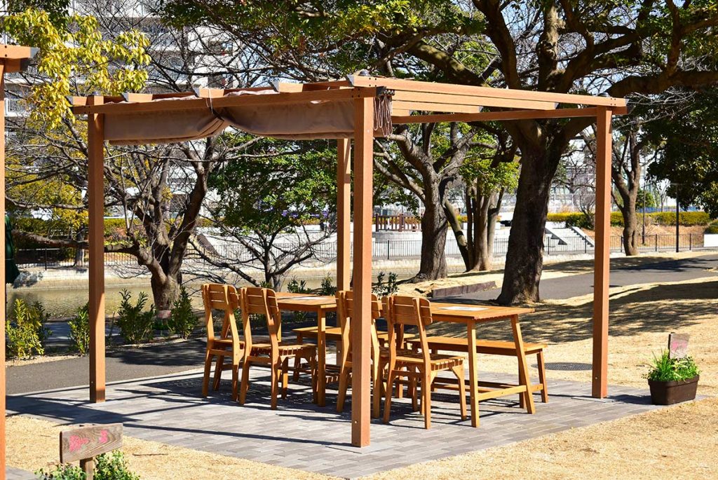 都内最新BBQ施設 THE RIVERSIDE BBQ NISHIKASAI - 川沿いに設置された木目調パーゴラのダイニングスペース。緑に囲まれた開放的な空間でラグジュアリーなBBQ体験を提供。DEPOS製3m×3mパーゴラ採用事例