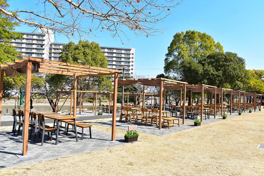 THE RIVERSIDE BBQ NISHIKASAI アウトドアダイニング - 河川敷に設置された木目調パーゴラ施設の全景。整然と並ぶDEPOSパーゴラと木製家具が作り出す上質なBBQ空間。東京都江戸川区の新設BBQ施設における、自然と都会的洗練さが融合したデザイン事例
