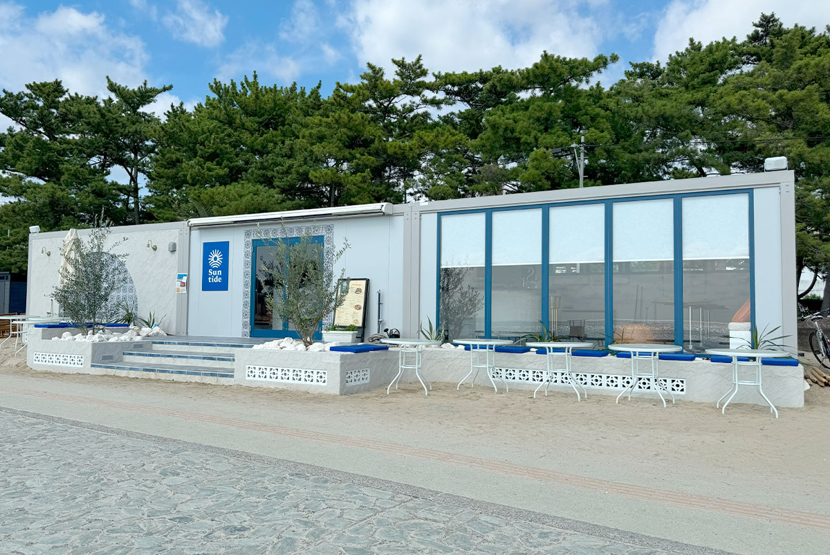 青空の下に建つSuntide Cafe、全面窓とオリーブの植栽がリゾート感を演出する外観風景